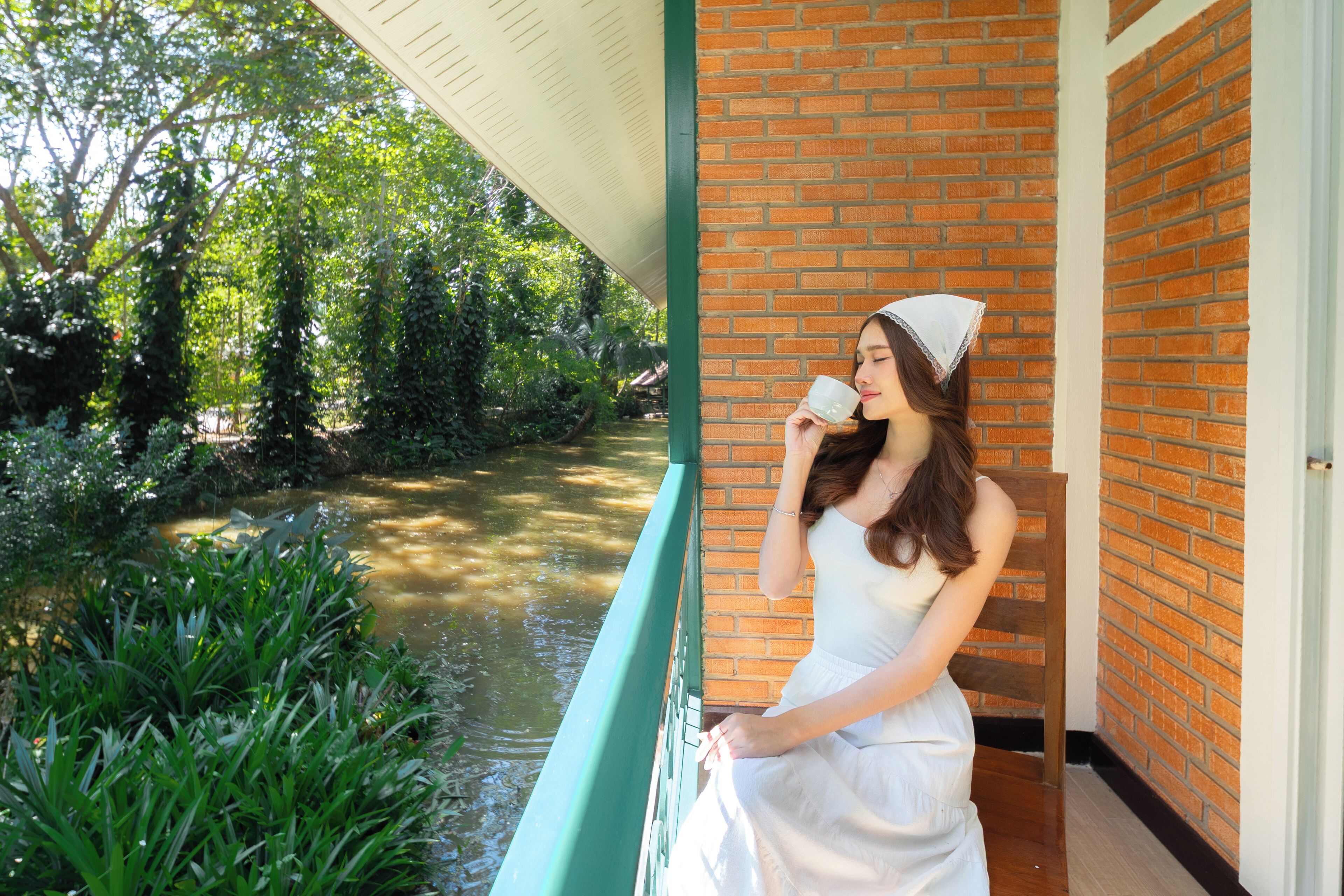 Une personne vêtue d’une robe blanche, appuyée sur une rambarde verte, surplombe un mur de briques et un canal d’eau bordé de végétation.