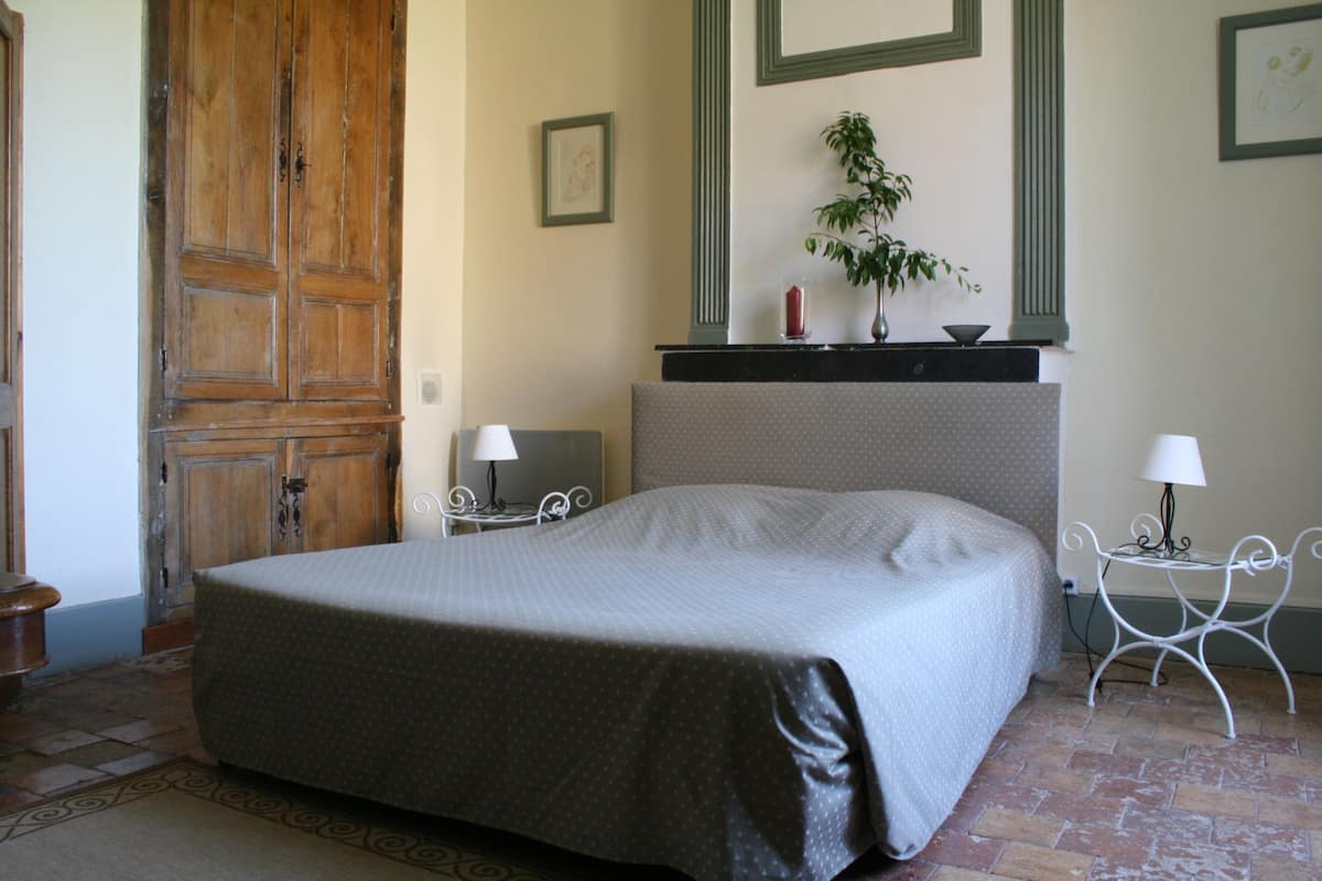 A bedroom with a large bed, a wooden wardrobe, a small table with a lamp, and a plant on a shelf.