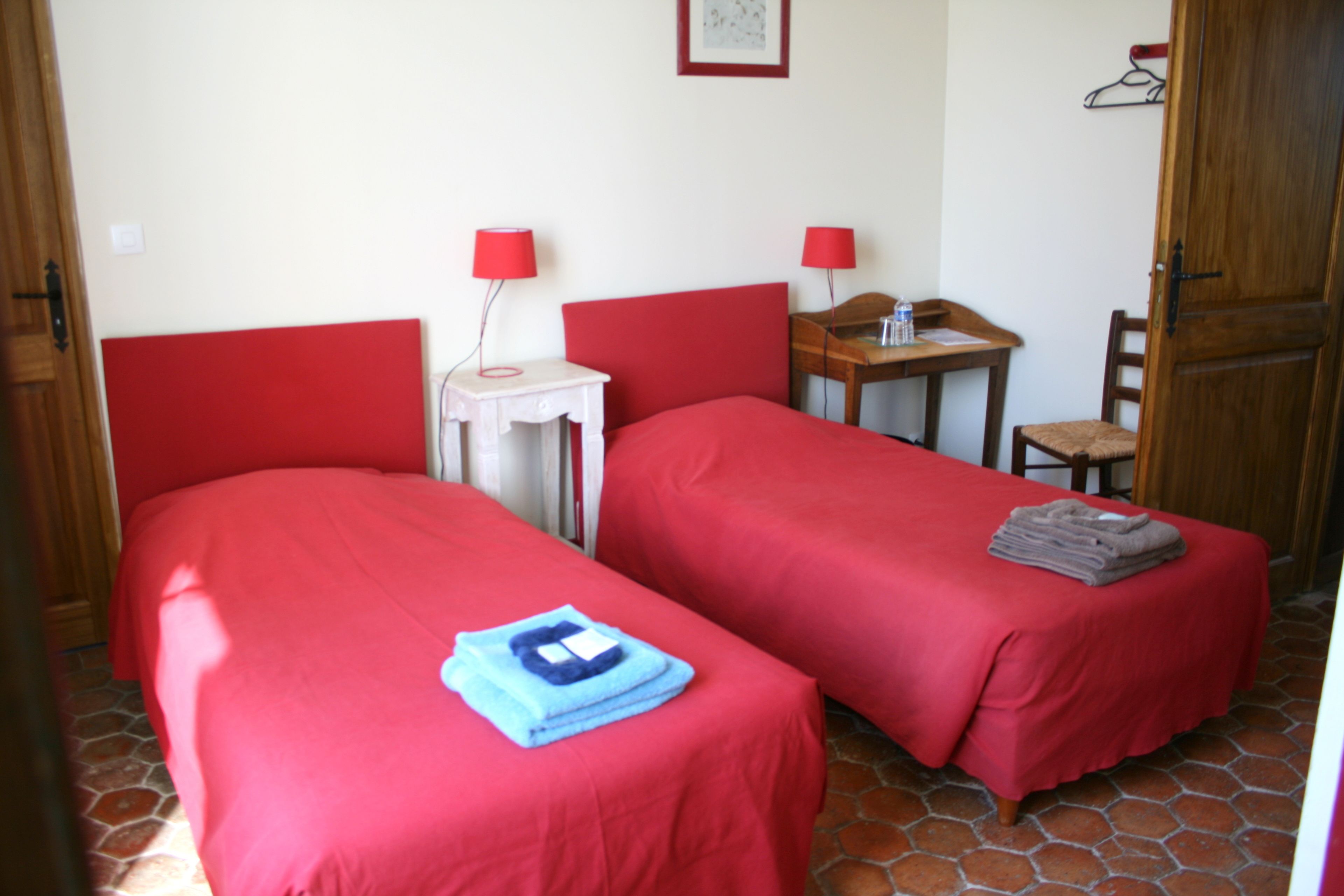 A room with two beds, each with a red bedspread and a blue towel on one bed. There is a small table with a red lamp and a chair.