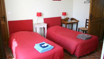 A room with two beds, each with a red bedspread and a blue towel on one bed. There is a small table with a red lamp and a chair.