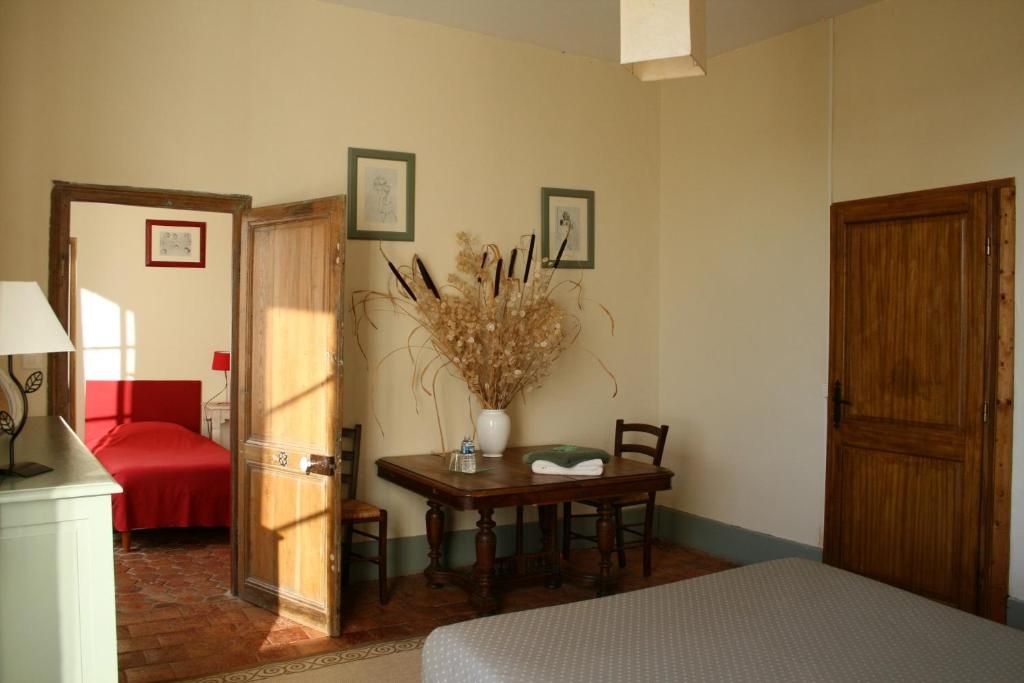 Una habitación con cama, una mesa con sillas, un florero con flores secas y una puerta.