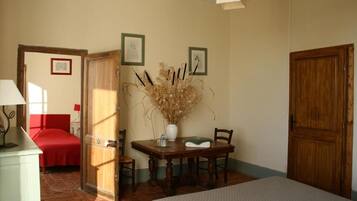 Una habitación con cama, una mesa con sillas, un florero con flores secas y una puerta.