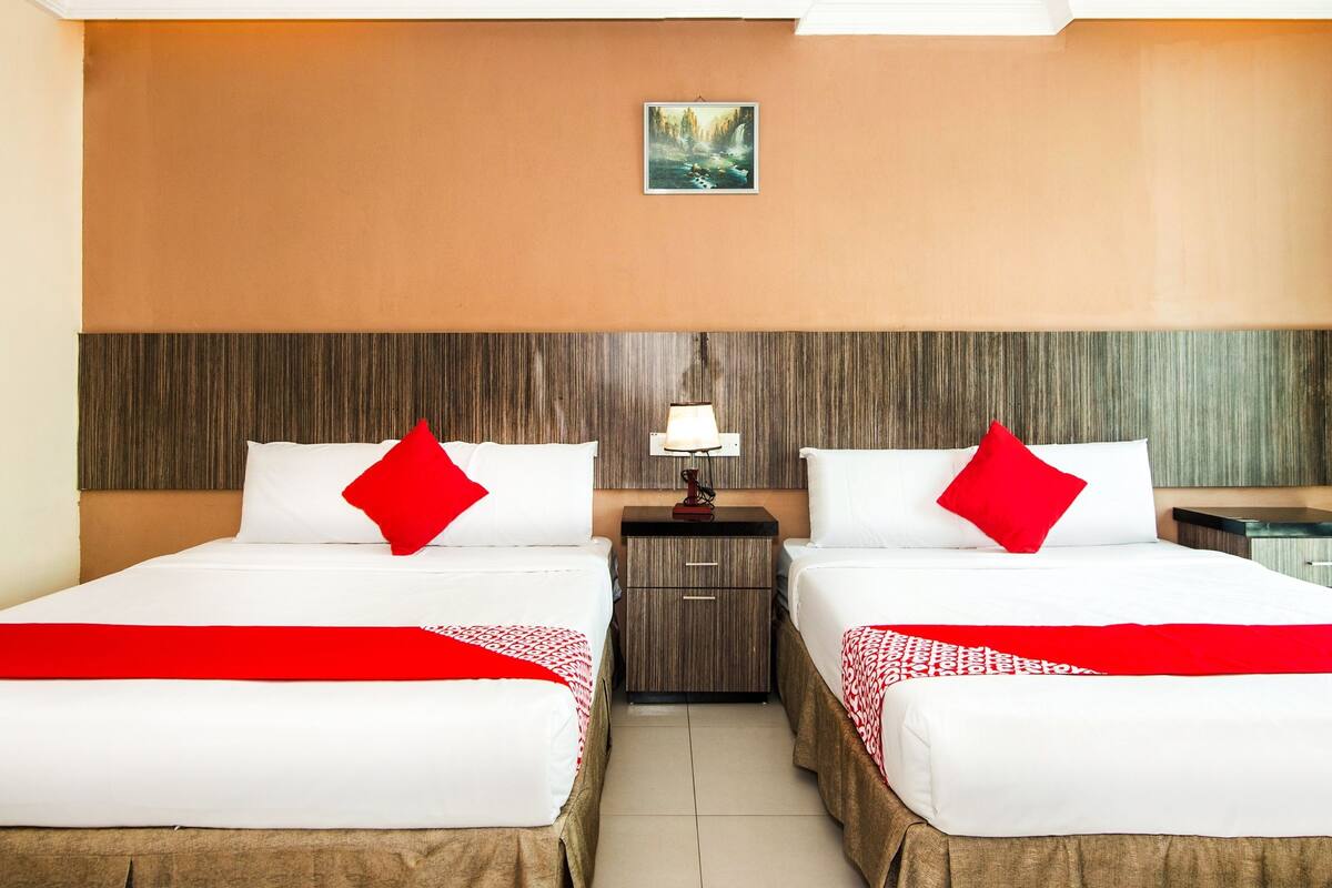 Habitación de hotel con dos camas, cada una con ropa blanca y almohadas rojas, una mesita de noche con una lámpara y un cuadro en la pared.