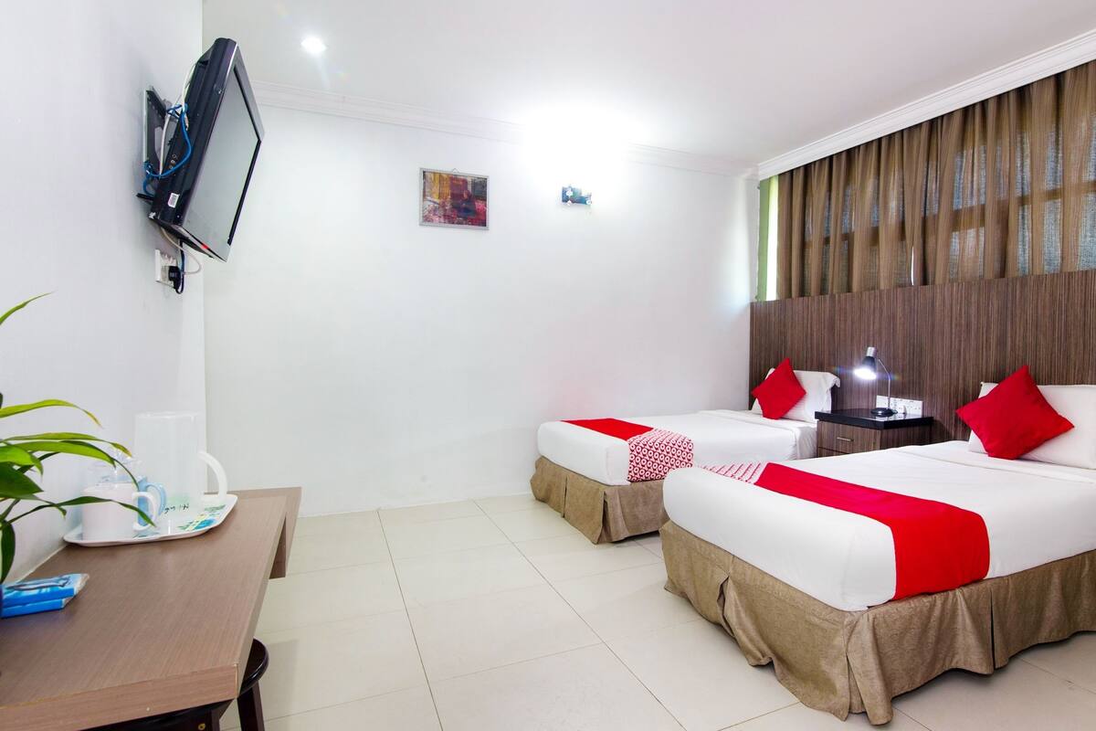 Habitación de hotel con dos camas, un televisor montado en la pared, un escritorio con una planta y una ventana con cortinas.