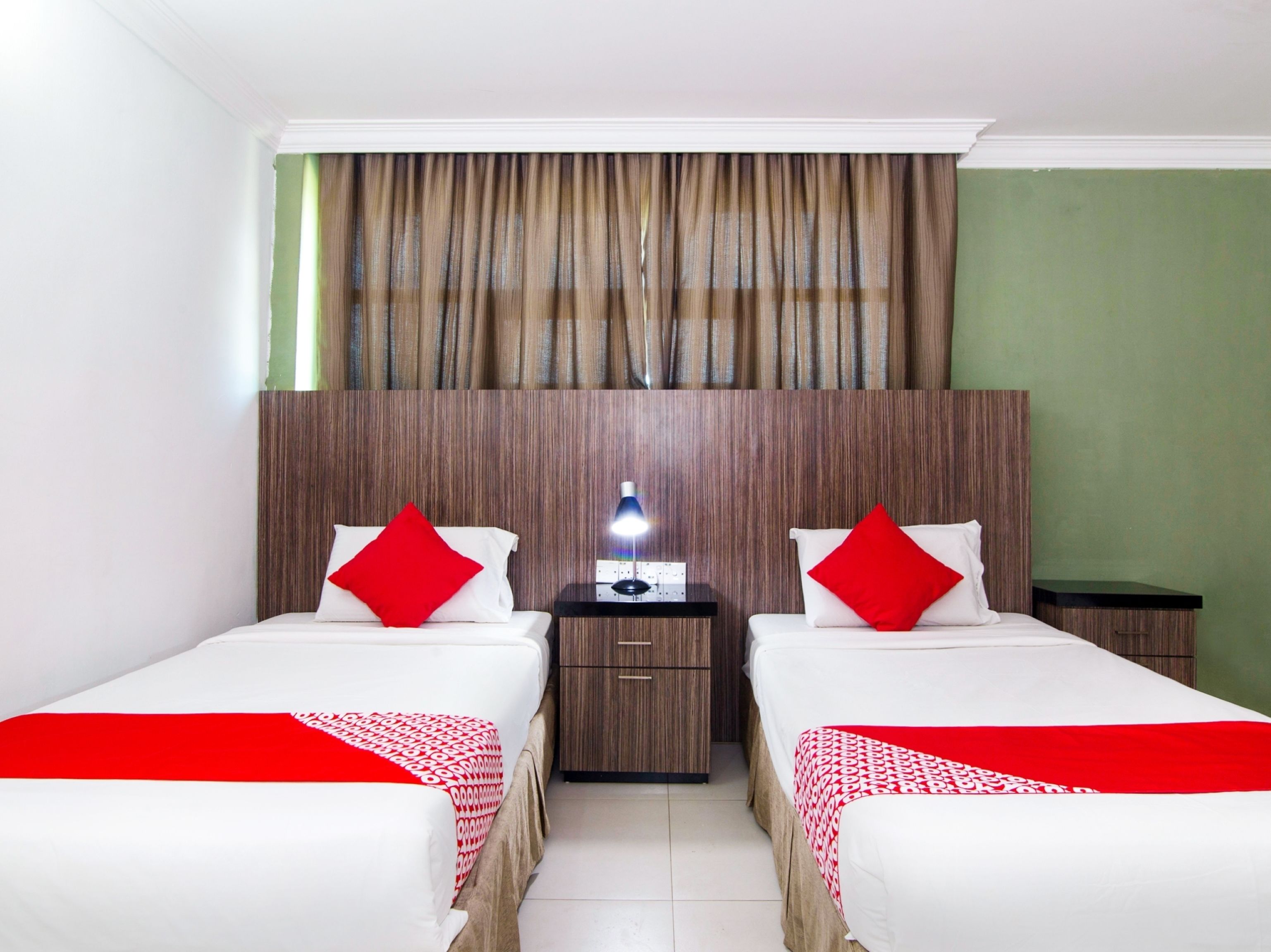 Une chambre d’hôtel avec deux lits, chacun agrémenté d’un oreiller rouge et d’une couvre-lit à motifs. Une table de chevet avec une lampe se trouve entre les deux lits.