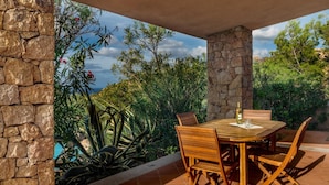 A stone patio with a wooden table and chairs, surrounded by lush greenery and a view of the sky. - Villa Agave (Trinità d'Agultu e Vignola)