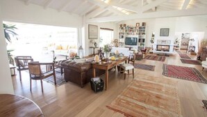 Un salon spacieux avec un grand canapé en cuir, une table basse en bois et une cheminée. On y trouve de nombreuses étagères remplies de livres et d’objets décoratifs. De grandes fenêtres permettent à la lumière naturelle d’entrer dans la pièce.