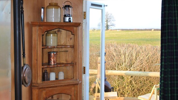 A wooden cabinet with jars and a lantern, a door to an outdoor area with a view of a field and a fence. - Glamping Thorpe (Banbury)