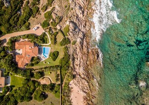 Vue aérienne d’une propriété située sur la côte, avec une piscine, un espace de stationnement et une plage rocheuse.