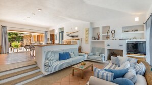 Ein geräumiges Wohnzimmer mit einer Sofagarnitur, einem Couchtisch und einem Fernseher. Im Hintergrund ist ein Kamin und eine Küchenzeile zu sehen.