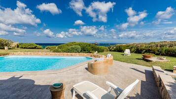 Piscina con vista al mar, rodeada de mobiliario exterior y vegetación.