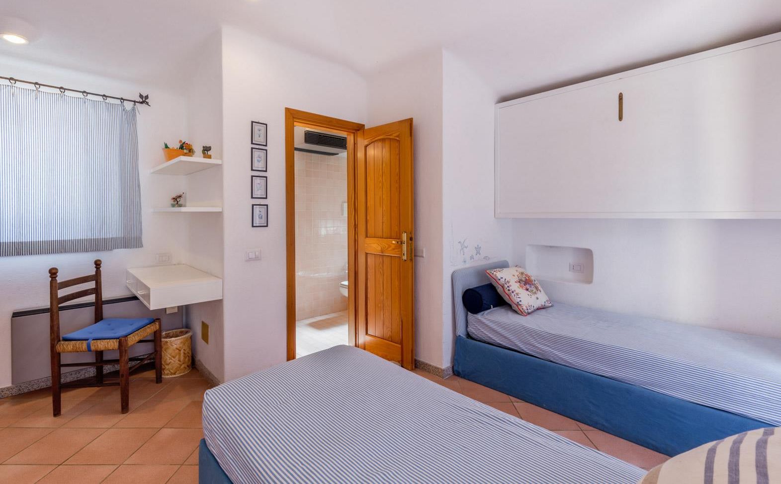 Un dormitorio pequeño y luminoso con dos camas individuales, una silla de madera, un escritorio y una ventana con cortinas.