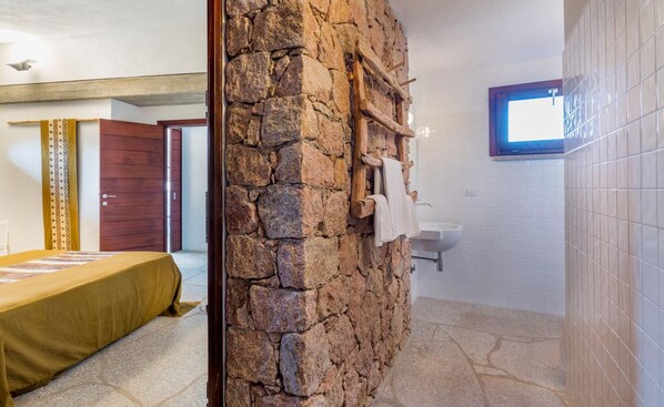 Una habitación con pared de piedra, una escalera de madera, una cama con colcha amarilla, una ventana y un baño con azulejos blancos.