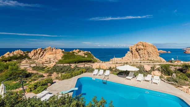 A pool with lounge chairs and umbrellas, surrounded by rocky terrain and greenery, with a view of the sea.