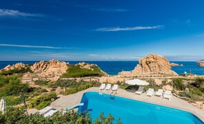 Una piscina con sillones de playa y sombrillas, rodeada de terreno rocoso y vegetación, con vista al mar.