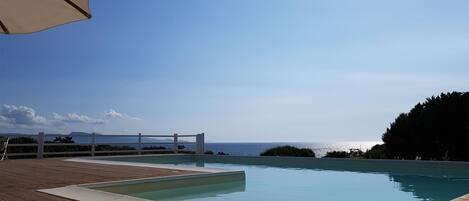 A wooden deck with a swimming pool, a white pergola, and a view of the ocean.