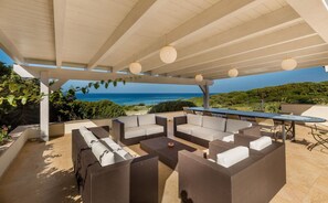 A covered outdoor seating area with a view of the sea, furnished with sofas and a table.
