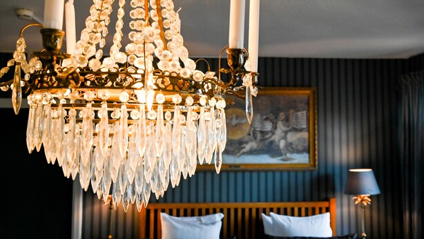 A chandelier with multiple candles, a bed with white pillows, and a painting on the wall. - Palace Hotell (Hultsfred)