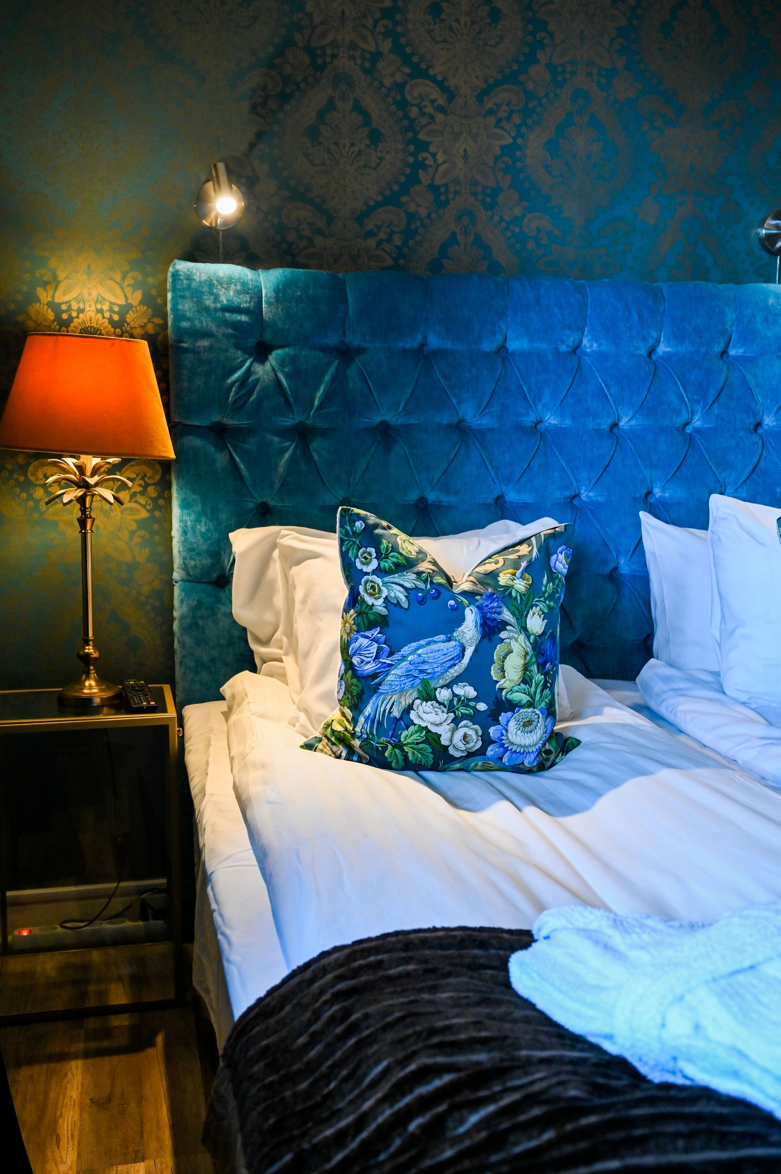 A bedroom with a tufted headboard, a floral pillow, white bedding, and a bedside lamp.