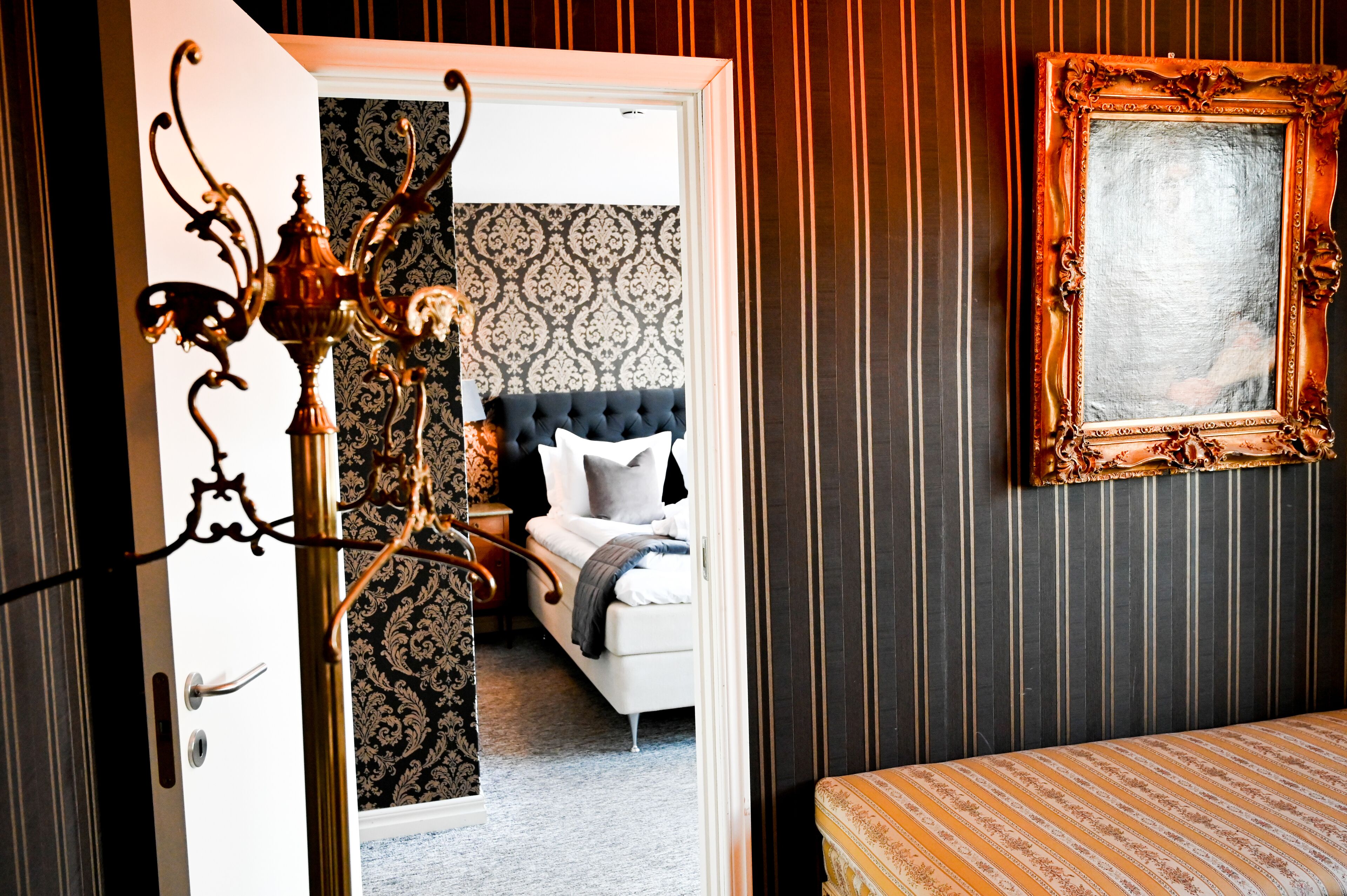 Une chambre à coucher avec un papier peint à motifs sombres, un lit doté d’une tête de lit capitonnée, un lustre et une œuvre d’art encadrée accrochée au mur.