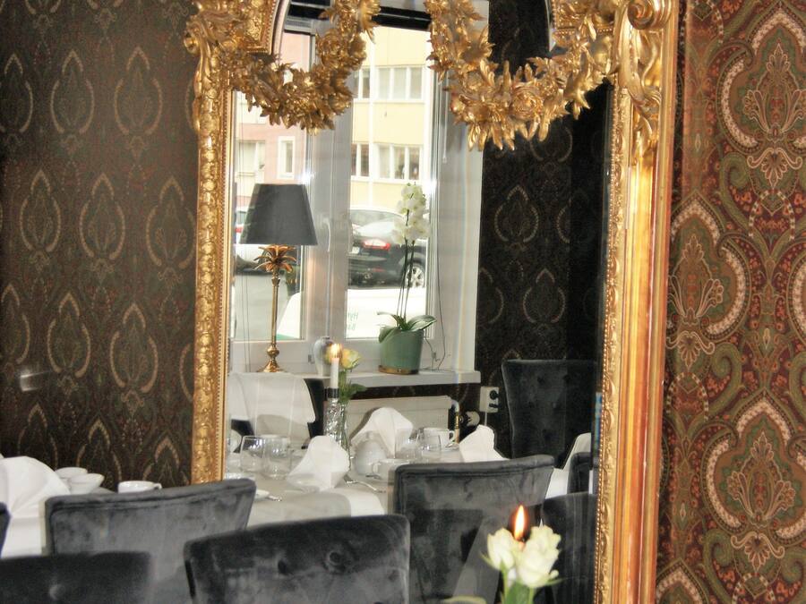 A dining area with a large ornate mirror, a table set for four with white tablecloths, black upholstered chairs, and a vase with white flowers.