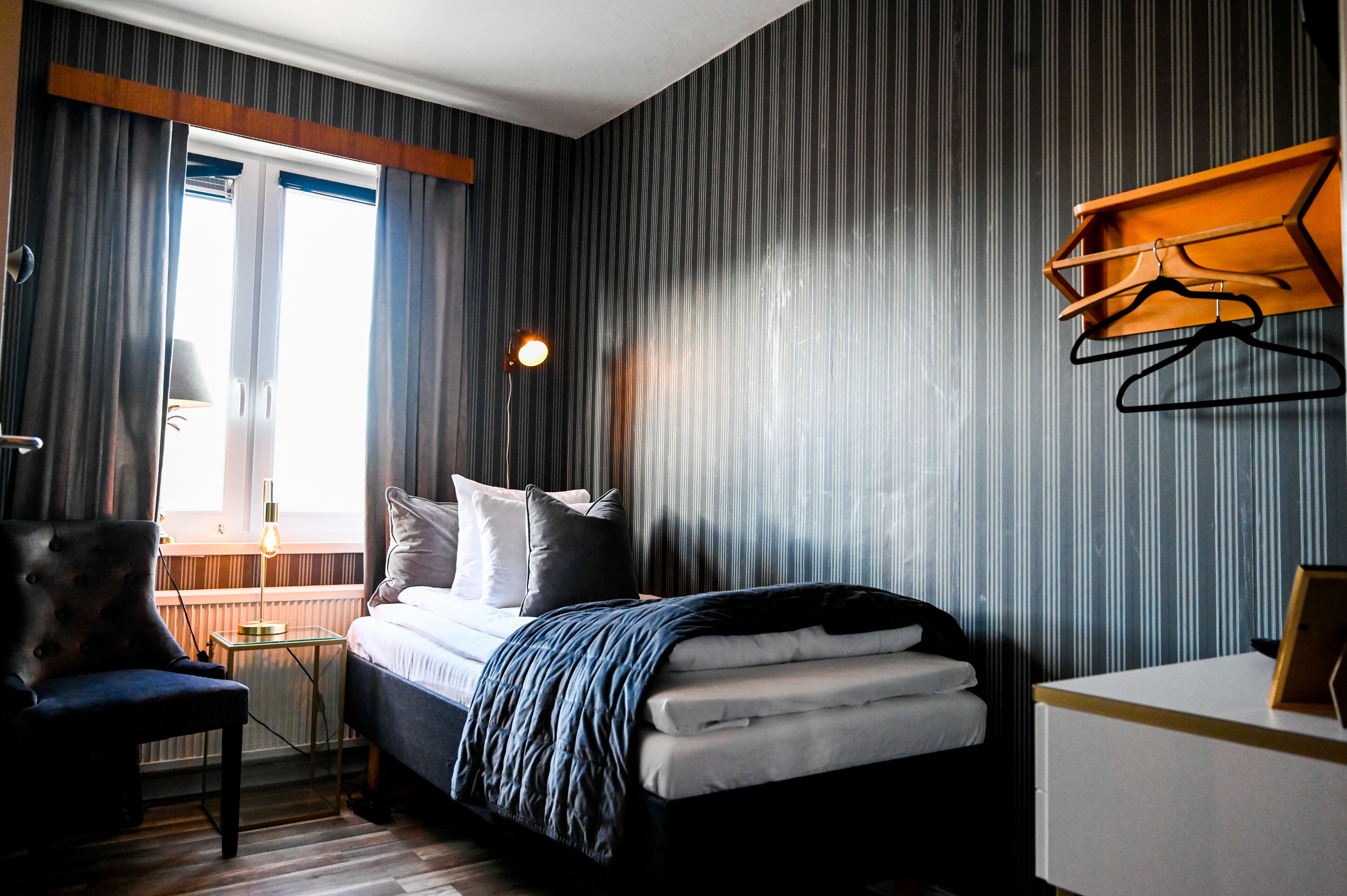 Une chambre d’hôtel avec un lit, une chaise, une table de chevet, une lampe et une fenêtre avec des rideaux.