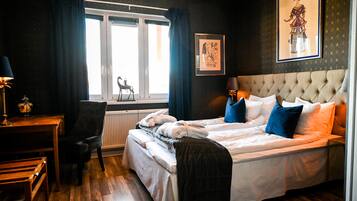 Habitación de hotel con cama, escritorio y silla. Hay una ventana con cortinas, un radiador y un cuadro en la pared.