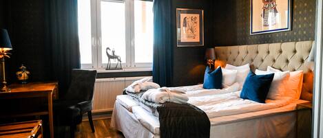 Habitación de hotel con cama, escritorio y silla. Hay una ventana con cortinas, un radiador y un cuadro en la pared.