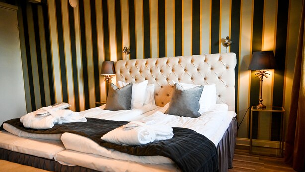 A hotel room with a large bed, a bedside table with a lamp, and a striped wall. - Palace Hotell (Hultsfred)
