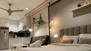 A modern bedroom with a large bed, bedside tables, a desk, and a kitchenette in the background.