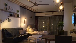 A modern living room with a sofa, coffee table, and dining table. There is a ceiling fan, pendant lights, and a TV mounted on the wall.