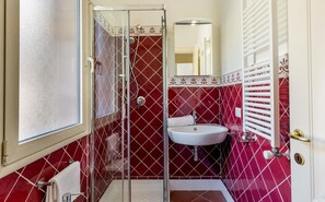 A bathroom with a glass shower enclosure, a white sink, and a red-tiled wall. - Villa Saluto del Sole (Trinità d'Agultu e Vignola)