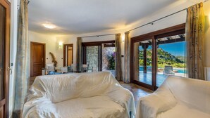 Un vaste espace de vie comprenant un canapé sous une pergola, une table à manger avec des chaises, et une grande porte-fenêtre coulissante donnant sur une piscine avec vue sur les collines.