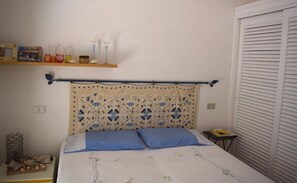 A bedroom with a bed, a headboard with a decorative pattern, a shelf with various items, and a window with blinds. - Villetta Ambra Rossa (Trinità d'Agultu e Vignola)