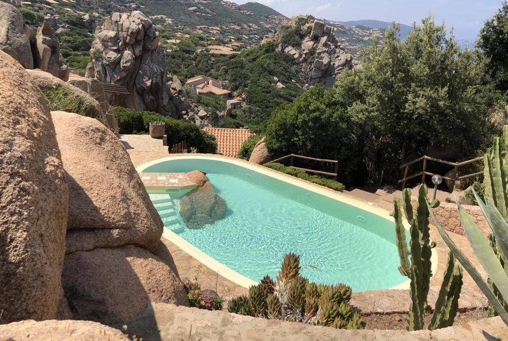 A pool surrounded by large rocks and cacti, with a view of a mountainous landscape in the background.
