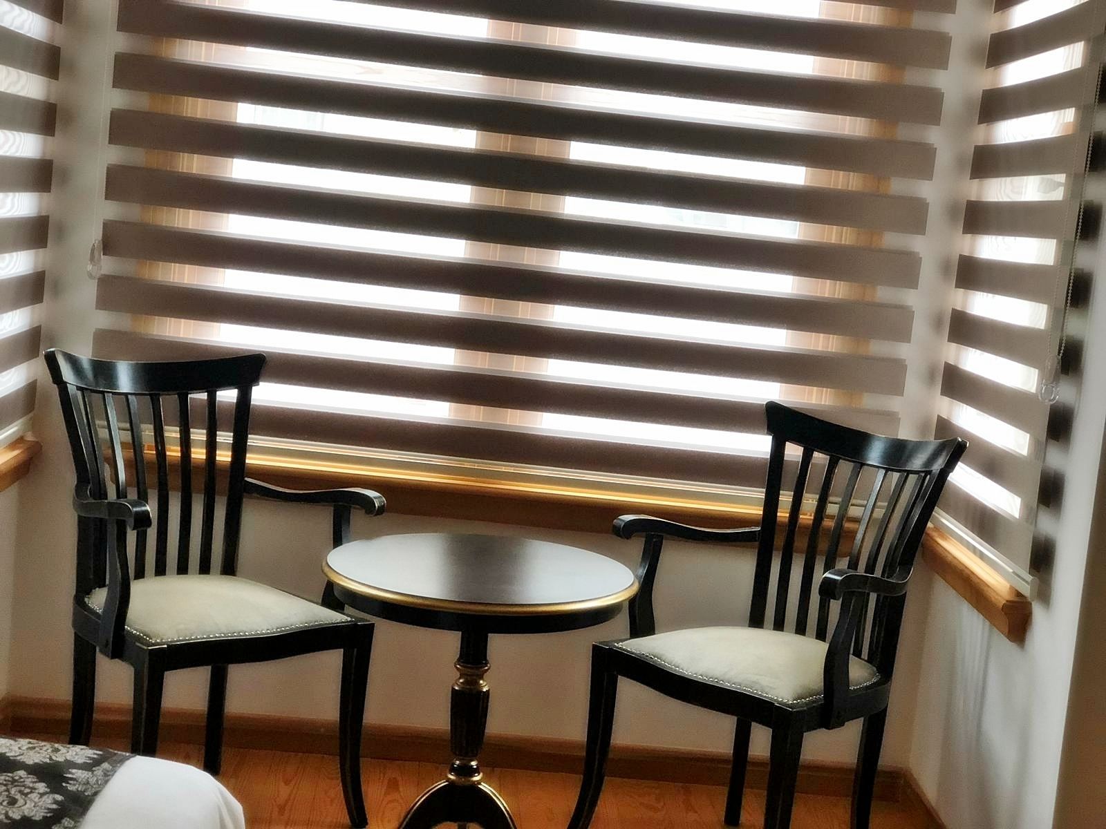 Eine Sitzecke mit zwei schwarzen Sesseln und einem kleinen runden Tisch, vor einem Fenster mit horizontalen Jalousien.