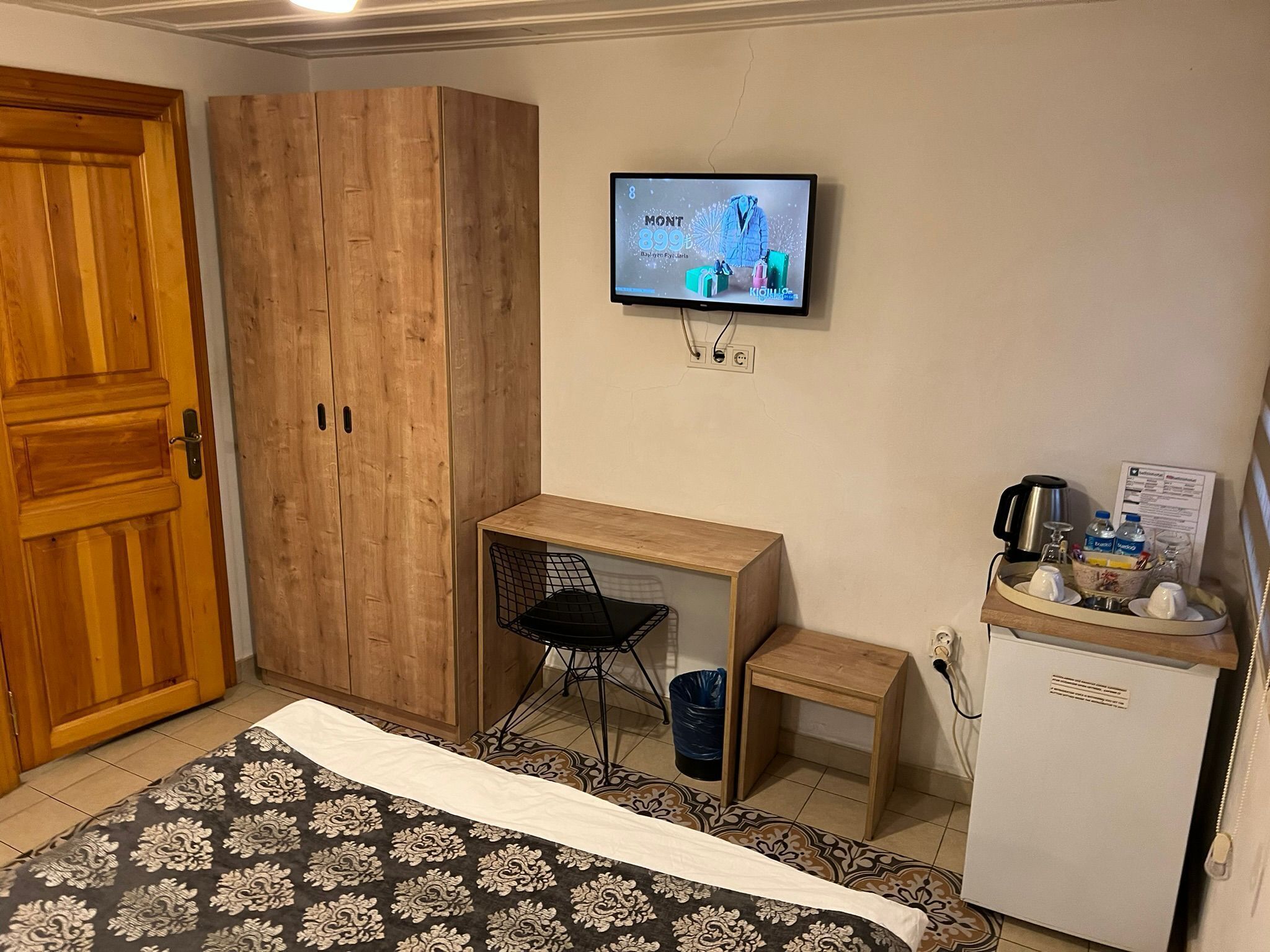 A bedroom with a bed, wooden wardrobe, desk, chair, television, mini-fridge, and a small table with a tray.