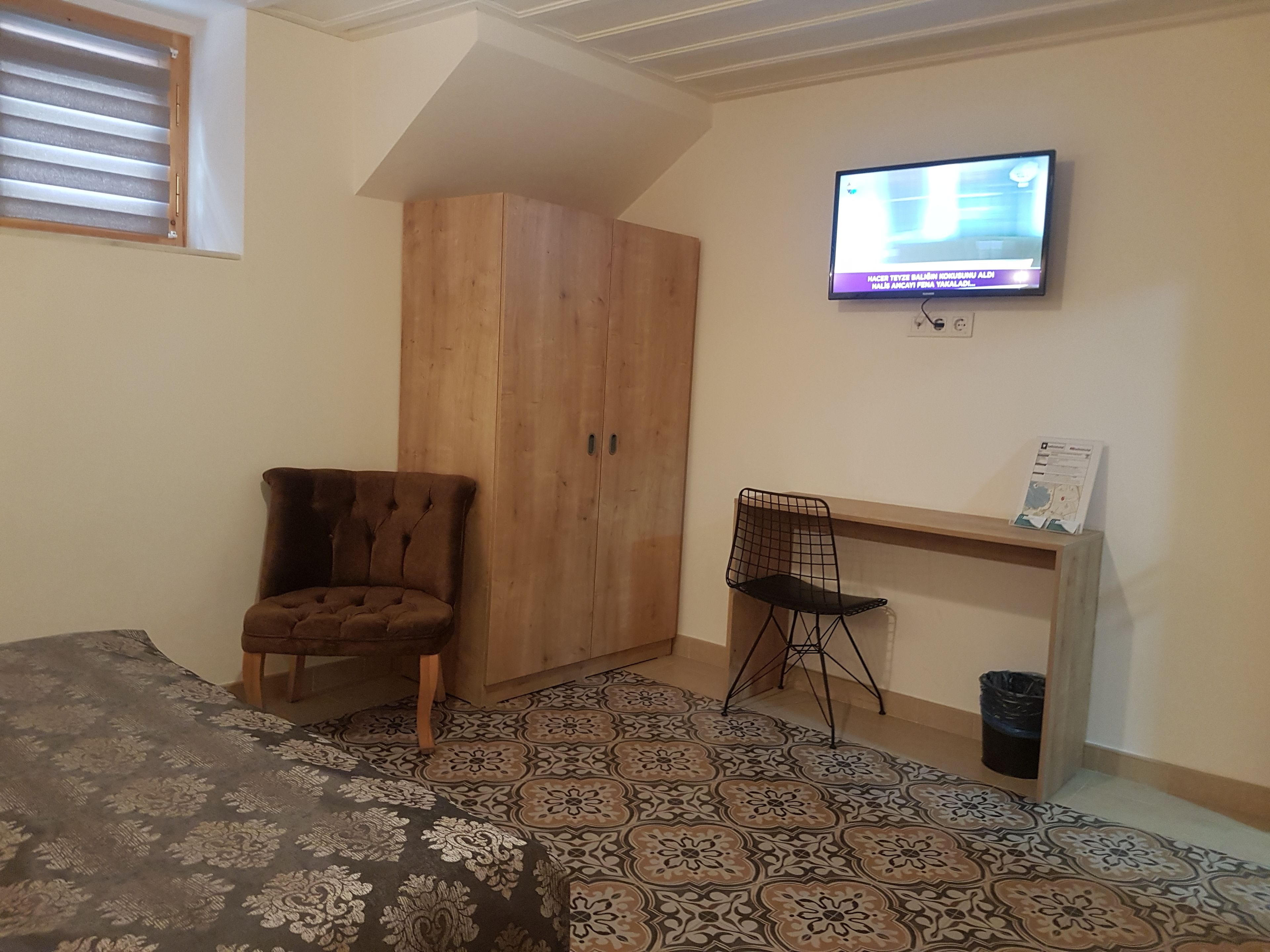 A bedroom with a bed, a wooden wardrobe, a desk with a chair, a television, and patterned floor tiles.