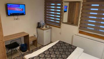 A hotel room with a bed, a desk, a television, a small refrigerator, and a window with blinds.