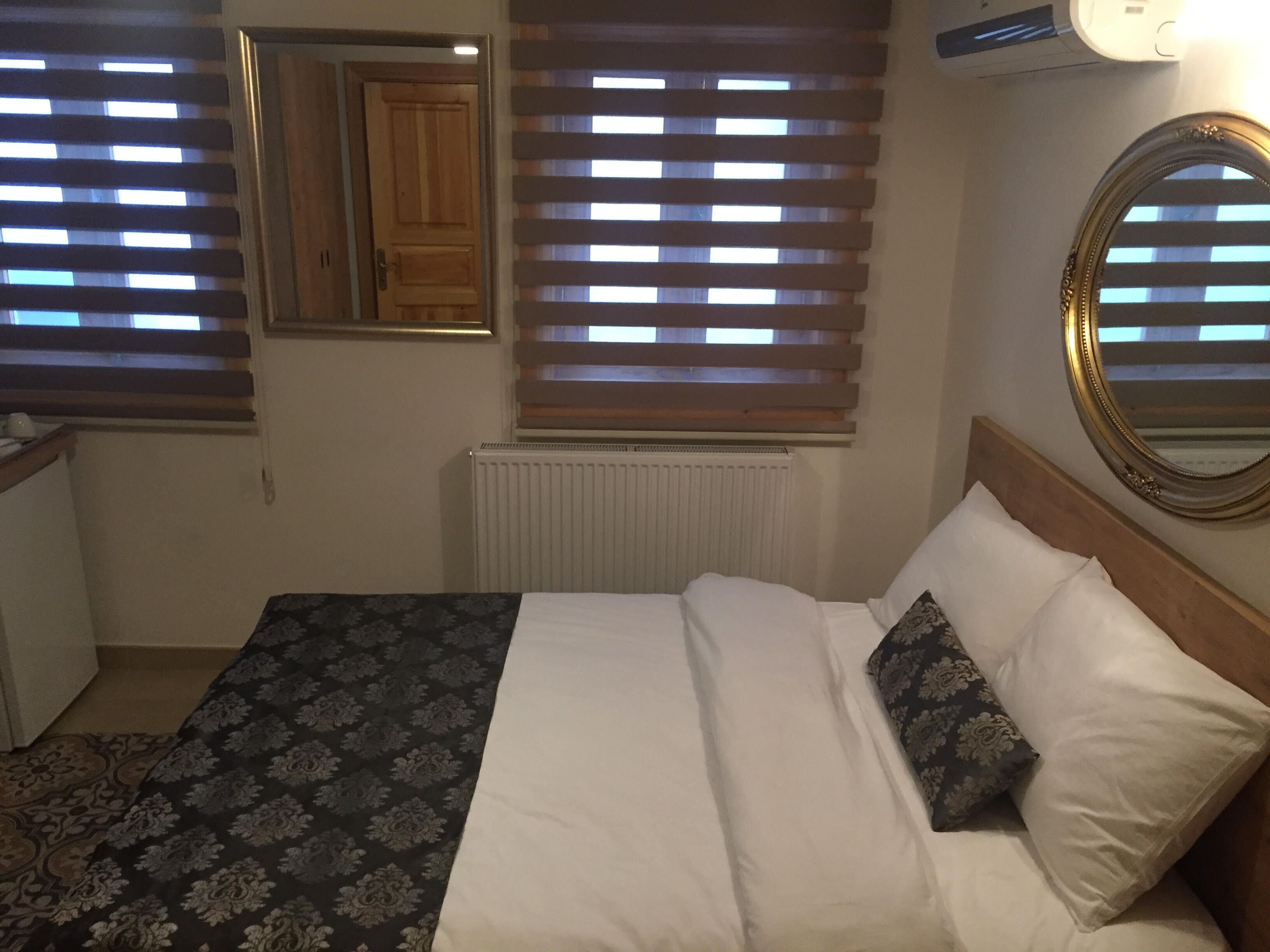 A bedroom with a bed, a radiator, a window with blinds, a mirror, and a wall-mounted light fixture.