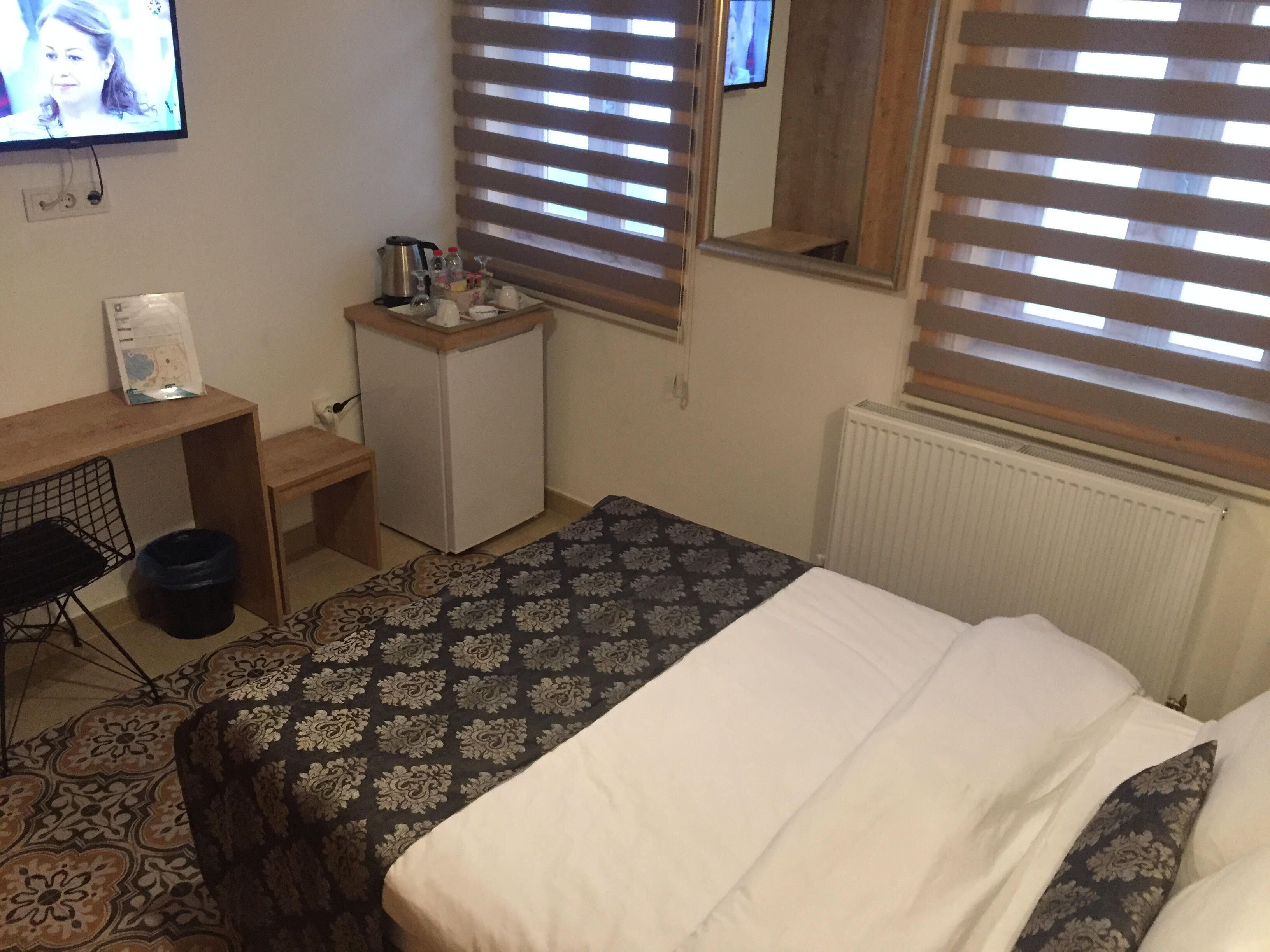 A bedroom with a bed, a desk, a small refrigerator, and a window with blinds.