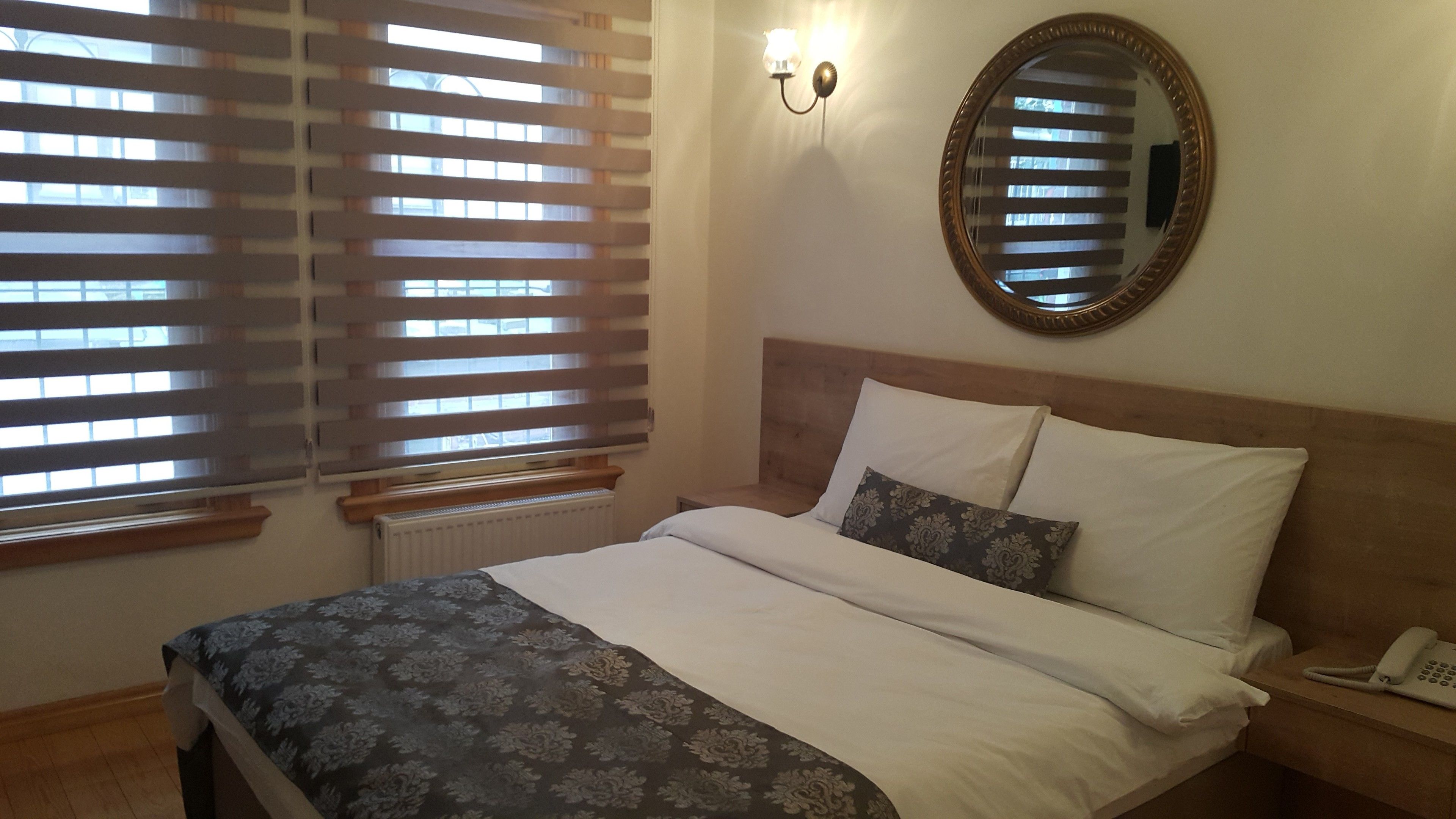 Een slaapkamer met een bed, nachtkastje, telefoon, een spiegel aan de muur en een raam met jaloezieën.