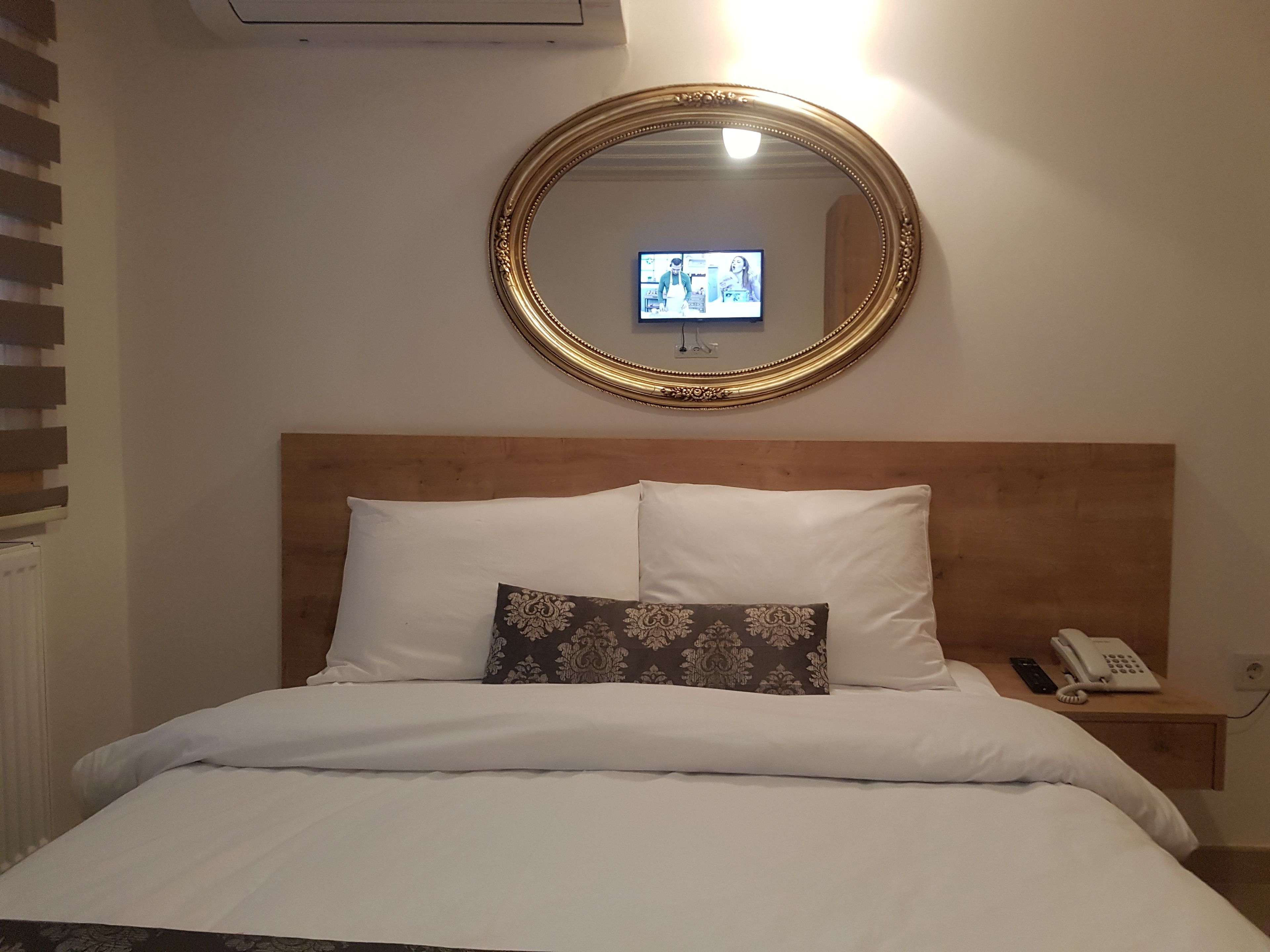 A neatly made bed with white bedding and a patterned pillow, a wooden headboard, a bedside table with a phone, and a mirror with a gold frame reflecting a television screen.