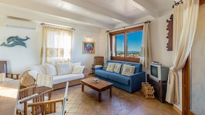 Ein Wohnzimmer mit einer Couch, einem blauen Sofa, einem Holztisch, Korbstühlen, einem Fernseher und Blick durch große Fenster.