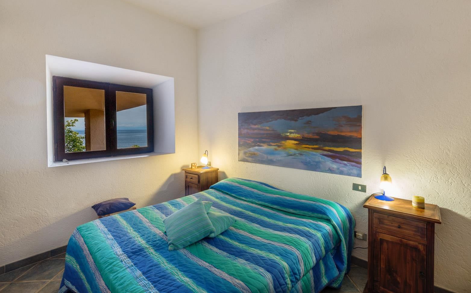 A bedroom with a bed, a nightstand, a lamp, a window with a view, and a painting on the wall.