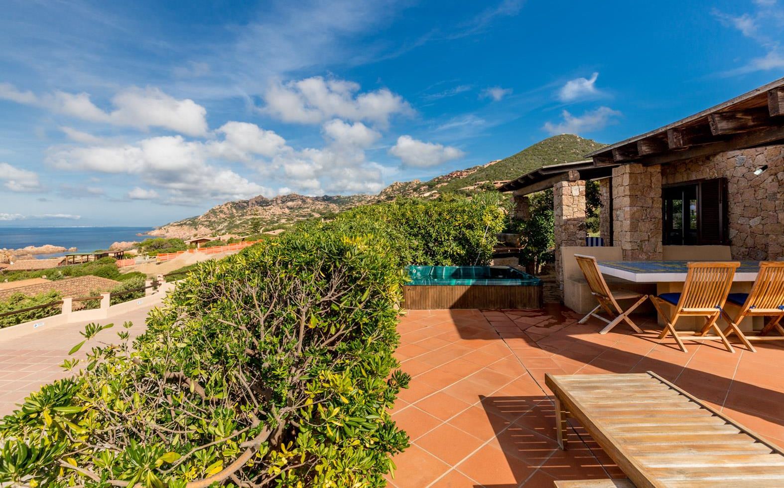 A patio with a table and chairs, a hot tub, and a view of the sea and hills.