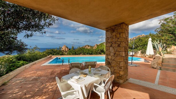 A covered outdoor dining area with a stone pillar, a pool, and a view of the sea and hills. - Villa Porcospino (Trinità d'Agultu e Vignola)