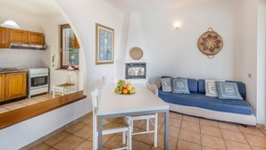 A compact kitchen with wooden cabinets, a dining table with a fruit bowl, a blue sofa with cushions, and a wall-mounted decorative item. - Villa Porcospino (Trinità d'Agultu e Vignola)
