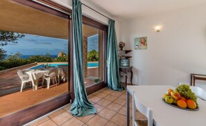 A room with a dining table and chairs, a view of a swimming pool, and a fruit bowl on the table. - Villa Porcospino (Trinità d'Agultu e Vignola)
