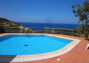 Une piscine rectangulaire à l’eau d’un bleu limpide, entourée d’une terrasse carrelée et d’une rambarde en métal. Au fond, on aperçoit la mer et un paysage vallonné.